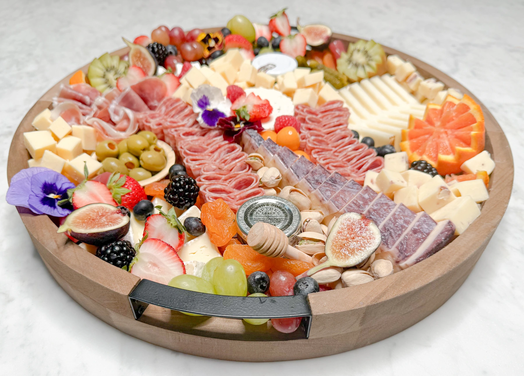 Fork and Flare medium charcuterie board at an angle on white marble in Orange County featuring Gouda, Manchego, Brie, Prosciutto, fresh fruit, nuts, and honey, handcrafted catering display.