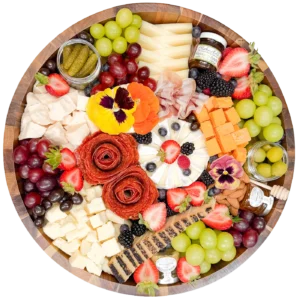 Round wooden charcuterie board loaded with cheeses, grapes, berries, sliced melon, meats, olives, crackers, jam, and edible flowers.