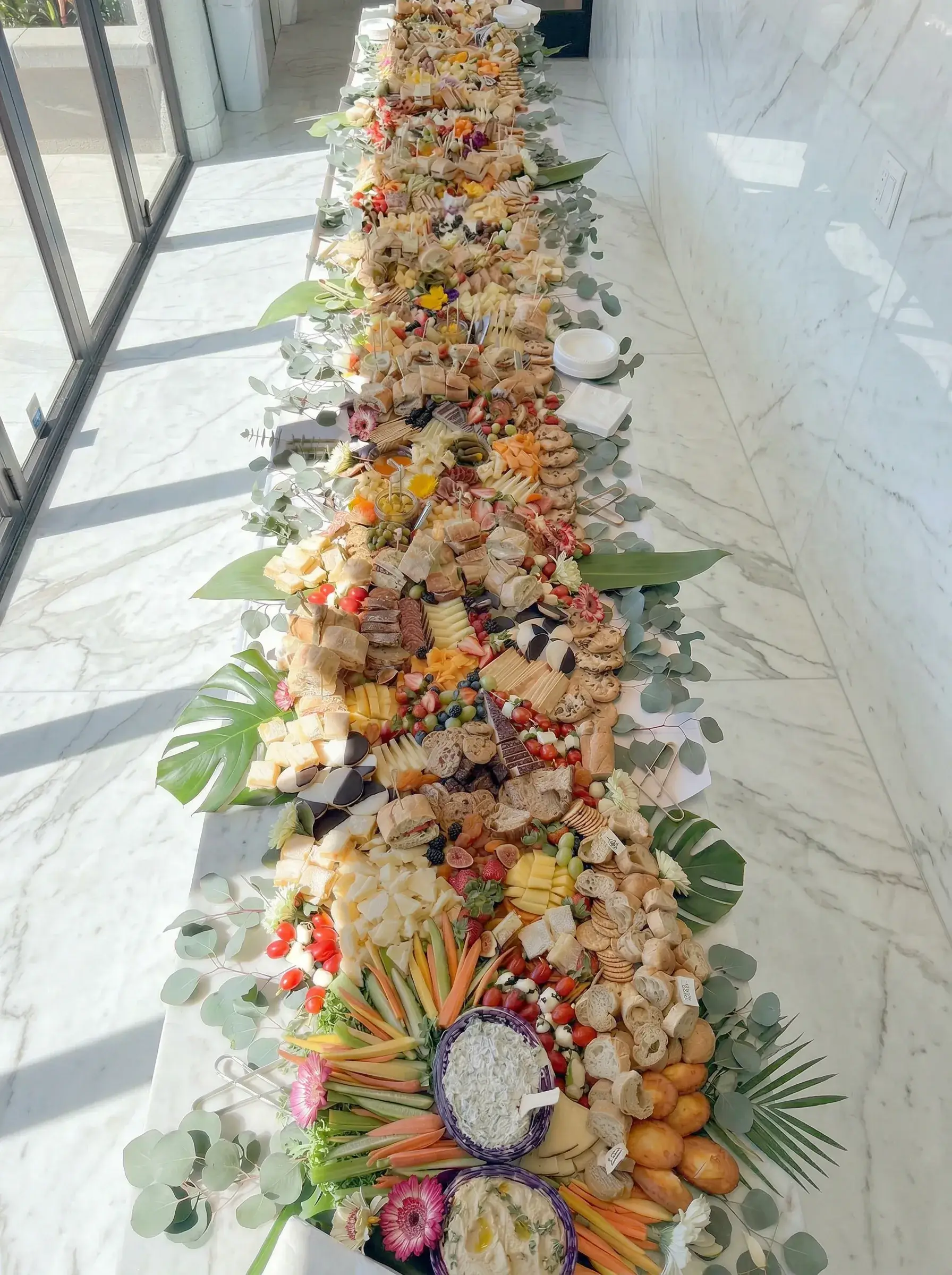 Flat lay grazing table by Fork and Flare in Mission Viejo, Orange County California designed for 150 guests featuring artisan cheeses, hand cut cured meats, fresh fruit, crudités, sandwiches, crackers, dips and floral greenery for luxury wedding and corporate event catering.