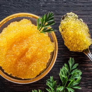 Golden caviar pearls in a glass bowl with a sprig of parsley on a dark wooden surface, plus a spoonful of caviar nearby